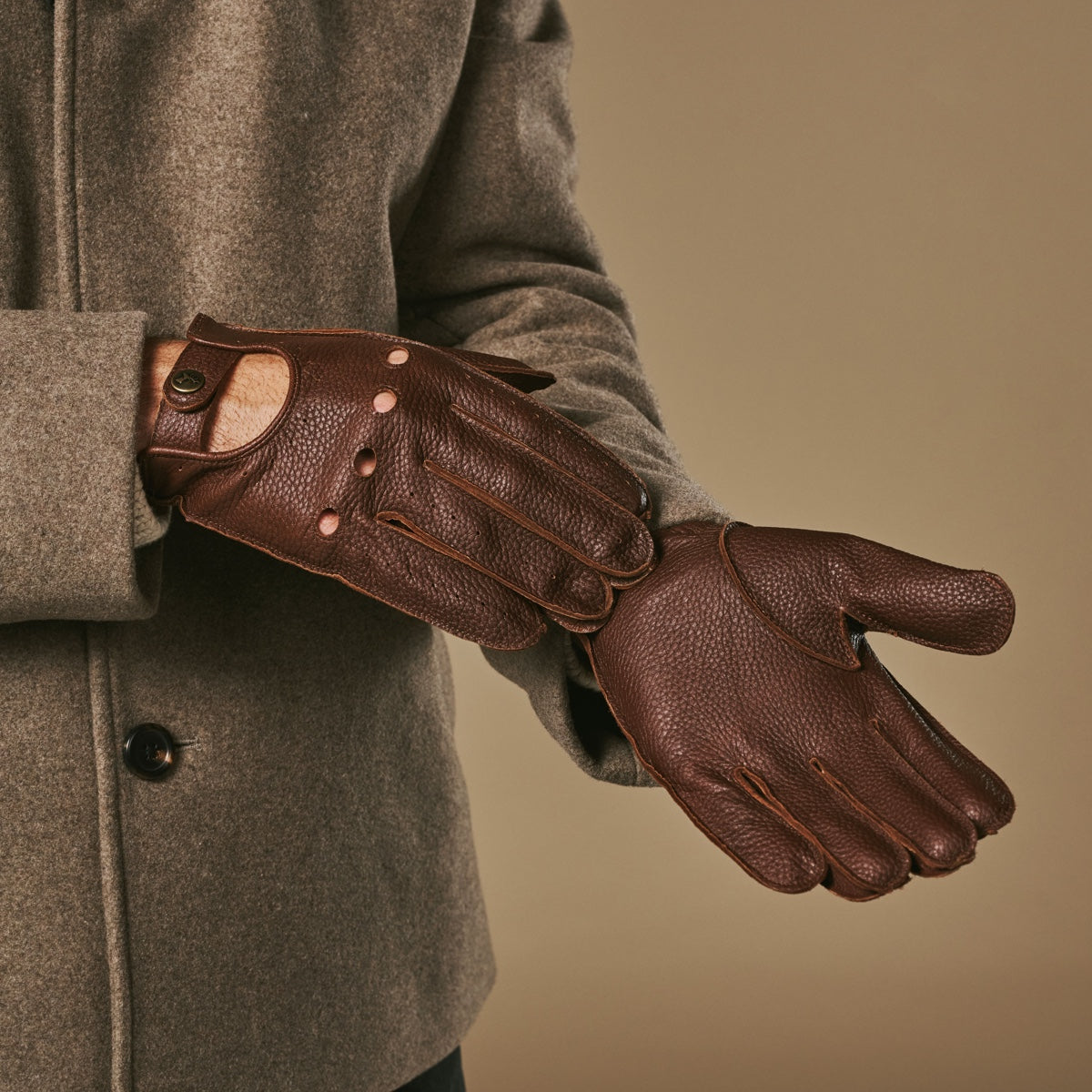 Brown deerskin driving gloves showing cuff strap detail and perforated fingertips.